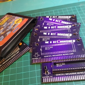 May include: A close-up of several black circuit boards with the text "The 8 Bit Manshed" printed on them. The boards are labeled "Genesis/Megdrive Repro Cart" and have a row of pins along the bottom edge. The boards are arranged in a stack, with the top board partially obscured by a black Sega Genesis game cartridge.