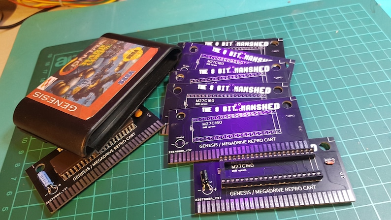 May include: A close-up of several black circuit boards with the text "The 8 Bit Manshed" printed on them. The boards are labeled "Genesis/Megdrive Repro Cart" and have a row of pins along the bottom edge. The boards are arranged in a stack, with the top board partially obscured by a black Sega Genesis game cartridge.
