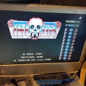 May include: A vintage arcade game screen displaying the title "TRUXION" with the game's copyright information and game stats. The screen is black with blue and white text and graphics. The game is set to round 1, stage 1.