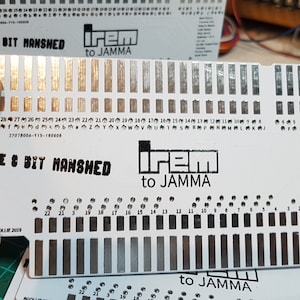 May include: A white card with black text that reads "The 8 Bit Manshed to JAMMA". The card has a grid of small black squares and a larger black square in the centre. The card is held in a hand.