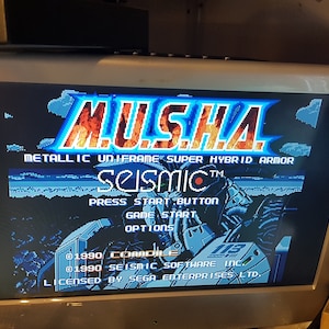 May include: A video game screen displaying the title "MUSHA" in large, stylized letters with a fiery orange and yellow background. The text "METALLIC UNIFRAME SUPER HYBRID ARMOR" is displayed below the title. The game's title screen also includes the text "SEISMIC&trade;" and the text "PRESS START BUTTON".