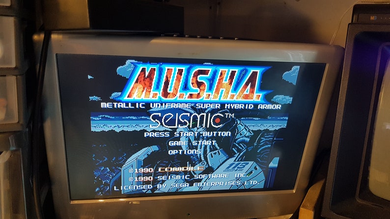 May include: A video game screen displaying the title "MUSHA" in large, stylized letters with a fiery orange and yellow background. The text "METALLIC UNIFRAME SUPER HYBRID ARMOR" is displayed below the title. The game's title screen also includes the text "SEISMIC&trade;" and the text "PRESS START BUTTON".