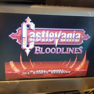 May include: A video game title screen for Castlevania: Bloodlines. The title is in a gothic font with a red and purple color scheme. The background is a red and orange landscape with a few spikes.
