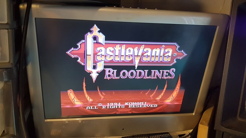 May include: A video game title screen for Castlevania: Bloodlines. The title is in a gothic font with a red and purple color scheme. The background is a red and orange landscape with a few spikes.