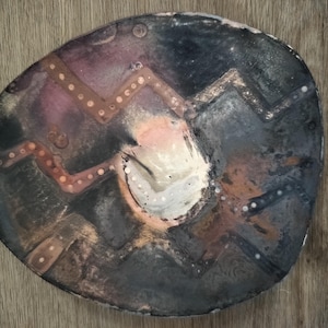 Może przedstawiać: Owalny ceramiczny talerz o ciemnej, nakrapianej powierzchni w odcieniach czerni, antracytu i głębokiego fioletu. Centralny, kremowy okrąg otoczony jest miedzianymi liniami w zygzak, ozdobionymi małymi, równomiernie rozmieszczonymi wypukłymi kropkami.