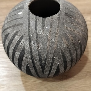 May include: A spherical dark gray vase with a rough, textured surface featuring deep, carved black geometric patterns. The vessel has a circular opening at the top and a rounded body, showcasing a contrast between the matte gray and black tones.
