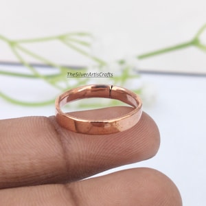 May include: A plain, polished copper ring with a smooth, flat band and a visible seam. The metal has a warm, reddish-brown metallic lustre. The text TheSilverArtisCrafts is printed in black above the ring in the background.
