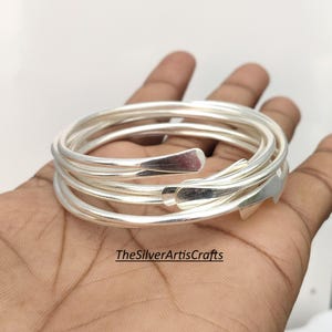 May include: A set of multiple stacked silver-colored bangles with a smooth, polished finish and tapered ends. The text TheSilverArtisCrafts is visible in the lower center of the image. The bangles are circular and metallic in appearance.