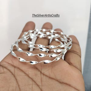 Può includere: Un set di tre braccialetti color argento caratterizzati da un design a spirale ritorto continuo. La superficie metallica è lucida e riflettente. Il testo TheSilverArtisCrafts è visibile in lettere nere sopra i braccialetti.