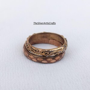 Könnte beinhalten: Ein Set aus zwei gestapelten Ringen mit metallischem Finish. Der obere Ring zeigt ein geprägtes Weinreben- und Blattmuster mit einer dunklen, gealterten Patina. Der untere Ring hat eine gehämmerte, texturierte Oberfläche. Text lautet TheSilverArtisCrafts.