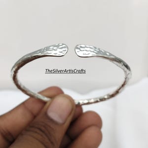 May include: A silver-colored open cuff bracelet featuring a textured, hammered surface throughout the band. The ends of the bracelet are flattened and rounded. The text TheSilverArtisCrafts is visible in the center of the open space.