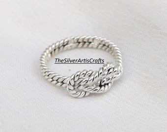 Puede incluir: Un anillo de plata con un diseño de cuerda trenzada que forma un nudo central. La banda está construida con dos hilos de plata entrelazados para crear una apariencia texturizada. El texto TheSilverArtsCrafts es visible en el anillo.