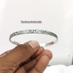 Pode incluir: Uma pulseira de prata fina com um padrão floral e de volutas em relevo na superfície externa. O metal tem um acabamento oxidado escuro nas áreas rebaixadas para destacar os detalhes. O texto TheSilverArtisCrafts é visível.