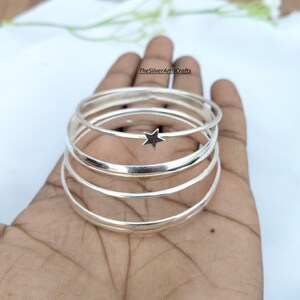 Peut inclure: Un ensemble de quatre bracelets joncs fins en métal argenté empilés. L'un des bracelets comporte une petite breloque en forme d'étoile à cinq branches. Le texte TheSilverArtisCrafts est visible en petites lettres noires en haut de la pile.