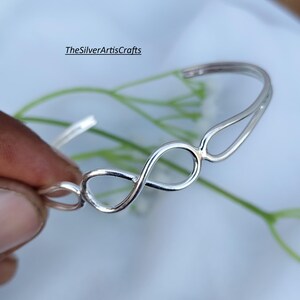 Puede incluir: Una pulsera de brazalete color plata que presenta un diseño central con el símbolo de infinito. La banda de metal tiene un acabado pulido y reflectante con una estructura abierta. El texto TheSilverArtisCrafts es visible en la imagen.