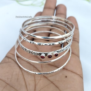 Puede incluir: Un conjunto de cinco brazaletes de plata con texturas y diseños variados. Uno de los brazaletes presenta tres pequeñas piedras granates redondas de color rojo intenso. Las superficies metálicas incluyen patrones lisos, grabados florales y de cuentas. El texto TheSilverArtisCrafts es visible.