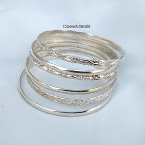 Op de afbeelding: Een stapel van vijf zilverkleurige armbanden met verschillende texturen, waaronder gladde, golvende en bloemmotiefbanden. De tekst TheSilverArtisCrafts is zichtbaar boven de sieraden. Elke armband heeft een cirkelvormige structuur.