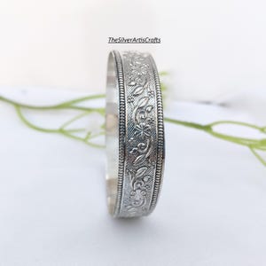 May include: A silver-toned bangle bracelet featuring an intricate embossed floral and vine pattern across its surface, bordered by beaded edges. The text TheSilverArtisCrafts is visible above the bracelet, which has a polished finish.