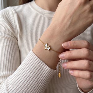 Puede incluir: Una pulsera de cadena dorada con un dije central en forma de perro estilizado, con una incrustación de nácar blanco y un pequeño cristal transparente. La pulsera incluye una cadena de eslabones finos y una pequeña etiqueta colgante.