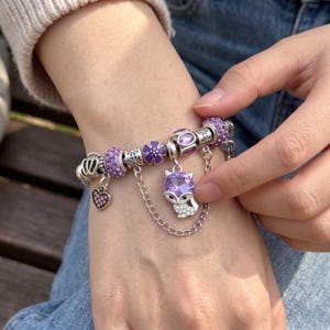Puede incluir: Una pulsera de dijes de plata con una variedad de cuentas moradas y plateadas, que incluye una flor morada, un dije en forma de corazón y un colgante central con forma de zorro con un gran cristal morado y una cadena de plata fina.