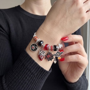 May include: A silver charm bracelet featuring various Halloween-themed charms including a red anatomical heart, a black spider web, a bat holding a pumpkin, a sun and moon disc, and faceted black and red beads on a metallic chain link band.