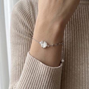 May include: A silver-toned chain bracelet featuring a central white, iridescent, flower-shaped charm with a small clear stone accent. The bracelet includes a mix of delicate link styles and a dangling charm at the end of the adjustable extension chain.