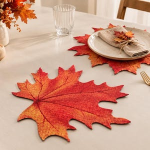 Op de afbeelding: Twee bladvormige placemats met een levendig kleurverloop van rood, oranje en geel. De ene placemat is leeg, terwijl op de andere een rond keramieken bord staat met een gevouwen beige servet, een kleine decoratieve herfsttak en een gouden vork.