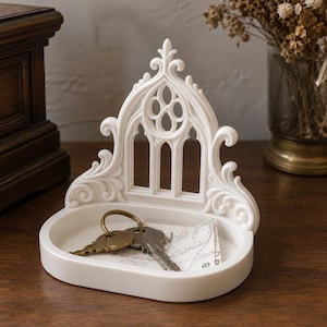 Puede incluir: Una bandeja de resina blanca con un panel trasero vertical que presenta un diseño de ventana arqueada y detalles de volutas. La bandeja tiene una base ovalada lisa que contiene dos llaves de metal y un pequeño papel con texto impreso.