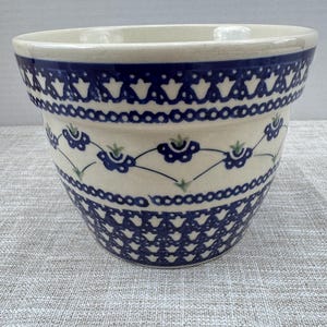 May include: A ceramic planter with a cream-colored base featuring a hand-painted dark blue pattern. The design includes a repeating geometric border at the top and bottom, with a central band of stylized blue flowers and green accents.