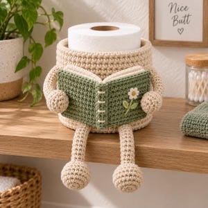 May include: A beige crochet toilet paper holder shaped like a figure with arms and legs, holding an open green crochet book with a small white flower detail on the cover. The holder is designed to sit on a flat surface with its legs dangling.