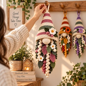 Peut inclure: Une décoration de gnome en crochet à suspendre avec un chapeau conique rayé crème, rose et vert, orné d'une fleur blanche et de baies. Le gnome possède un nez couleur crème et une longue barbe texturée faite de feuilles et de fleurs crochetées.