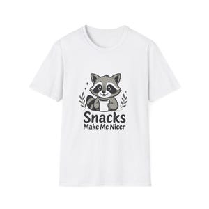 May include: A white short-sleeved t-shirt featuring a graphic of a grey raccoon holding a white cup. The raccoon is surrounded by small leaf sprigs and sparkles. Below the illustration, the text reads "Snacks Make Me Nicer" in black.