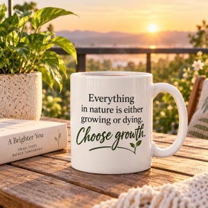 May include: A white ceramic mug featuring the black printed text: Everything in nature is either growing or dying. Choose growth. The mug has a curved handle and is positioned on a wooden surface next to a book titled A Brighter You.