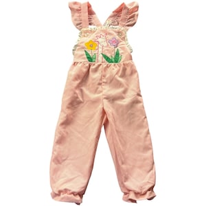 May include: A light pink corduroy overall featuring ruffled shoulder straps and a central embroidered applique of a giraffe surrounded by a yellow flower and a purple flower with green leaves. The garment has a gathered waist and cuffs.