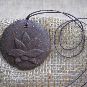 May include: A brown ceramic pendant with a raised flower design, hanging from a brown cord.