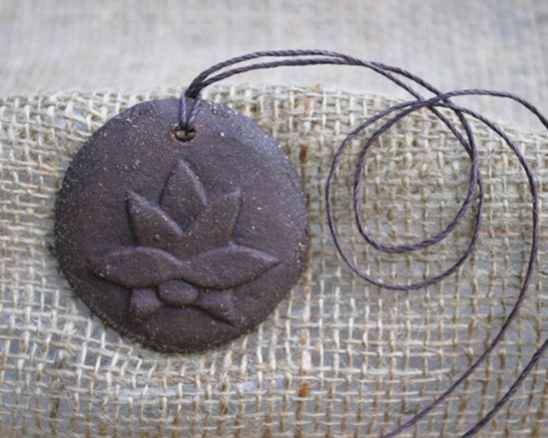 May include: A brown ceramic pendant with a raised flower design, hanging from a brown cord.