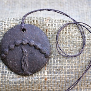 May include: A brown ceramic pendant with a tree design. The pendant is hanging from a brown cord.