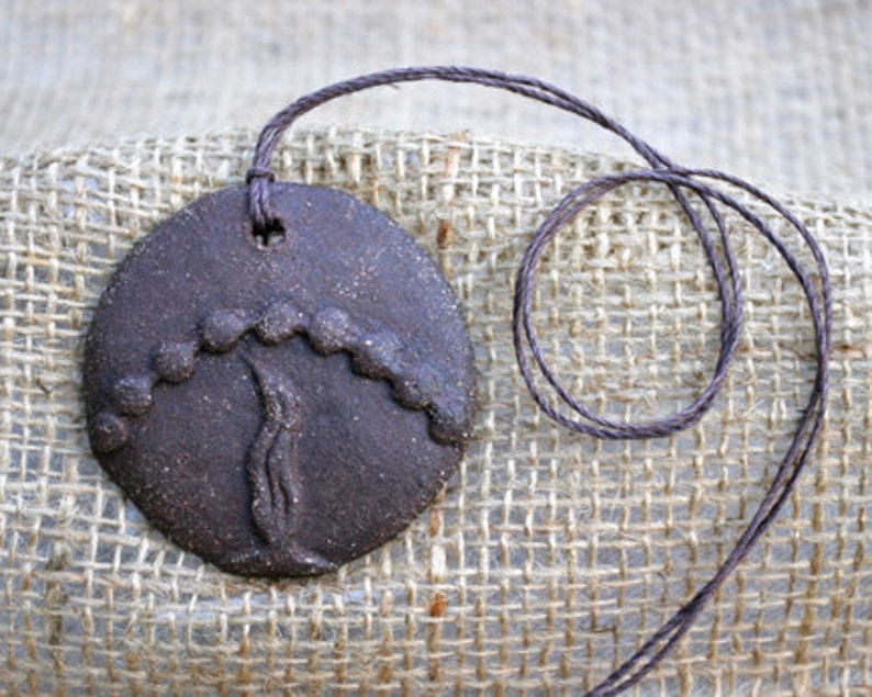 May include: A brown ceramic pendant with a tree design. The pendant is hanging from a brown cord.