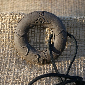 May include: A dark gray ceramic ring pendant with intricate carvings and a black leather cord. The pendant features a circular design with a hole in the center and various symbols carved into the surface.