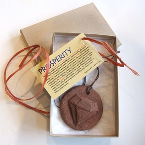 May include: A brown clay pendant with a diamond design, hanging from a brown cord in a white box with a brown paper tag that reads "PROSPERITY" and explains the symbolism of the pendant.