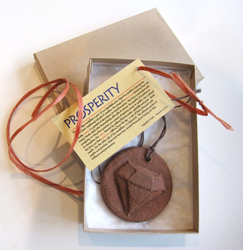May include: A brown clay pendant with a diamond design, hanging from a brown cord in a white box with a brown paper tag that reads "PROSPERITY" and explains the symbolism of the pendant.