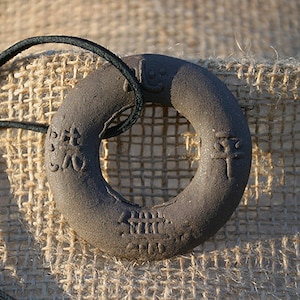 May include: A dark gray ceramic ring with Chinese characters inscribed on it. The ring is strung on a black cord and is lying on a burlap surface.