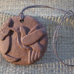 May include: A brown clay pendant with a bird design, hanging from a brown cord.
