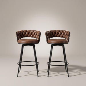 Puede incluir: Dos taburetes de bar con asientos y respaldos de cuero marrón con un diseño trenzado y acolchado. Cada taburete tiene una estructura de metal negro con cuatro patas delgadas y un anillo reposapiés circular como base del mecanismo giratorio.