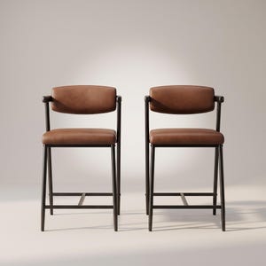 Puede incluir: Dos taburetes de bar con estructuras de metal marrón oscuro y tapicería de cuero liso color marrón medio en los asientos y respaldos curvos. Cada taburete tiene cuatro patas rectas conectadas por barras de soporte y pequeños apoyabrazos.