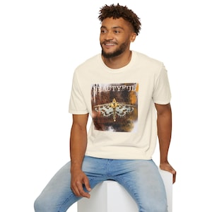 Op de afbeelding: Een crèmekleurig T-shirt met korte mouwen en een grafische print van een mot met gele, zwarte en witte vleugels. Boven de mot staat het woord BEAUTYFUL in een wit, verweerd schreeflettertype op een donkere, getextureerde achtergrond.