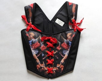 May include: Black corset with red ribbon lacing and a print of a painting featuring two angels. The corset has a V-neck and adjustable straps. The label reads "RetroFolie".