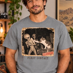 May include: A grey short-sleeve t-shirt featuring a graphic print of a cat interacting with two aliens. The image has a distressed border, a red stamp reading CLASSIFIED, and the text FIRST CONTACT printed in black below the graphic design.