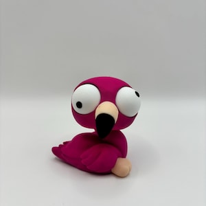 Puede incluir: Una pequeña figura de flamenco color magenta con una cabeza de gran tamaño y ojos grandes, redondos y blancos con pupilas negras. El ave tiene un pico de color beige claro con la punta negra y un pequeño pie beige visible al frente.