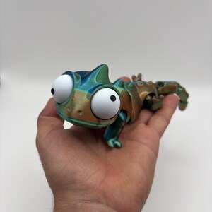 Peut inclure: Une figurine de caméléon articulée imprimée en 3D avec une finition métallique aux reflets changeants dans des tons bleu, vert et bronze. La créature possède de grands yeux blancs ronds avec des pupilles noires et un corps segmenté articulé.